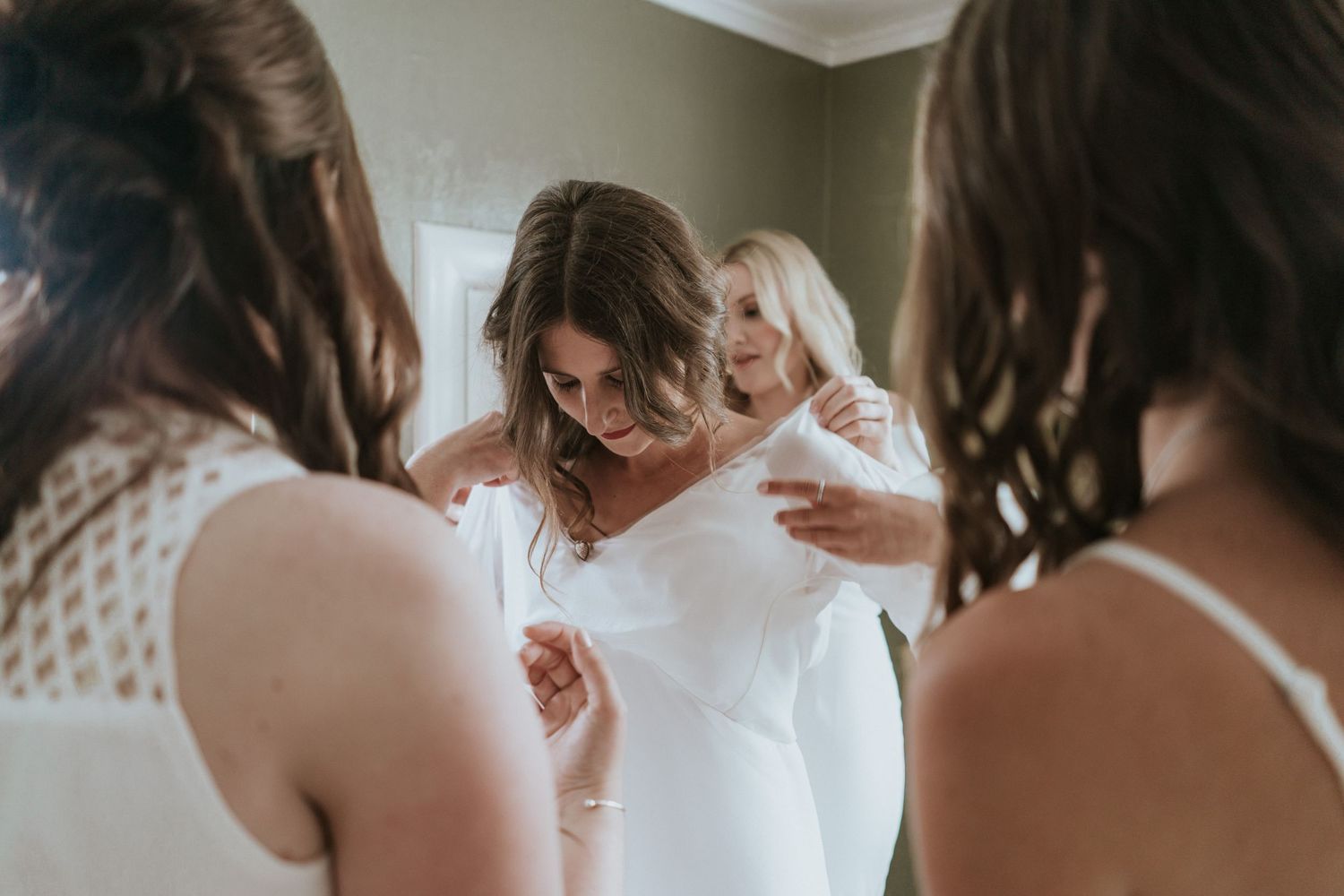 A bride in a white gown prepares for her wedding day while being helped by attendants in matching blush dresses.