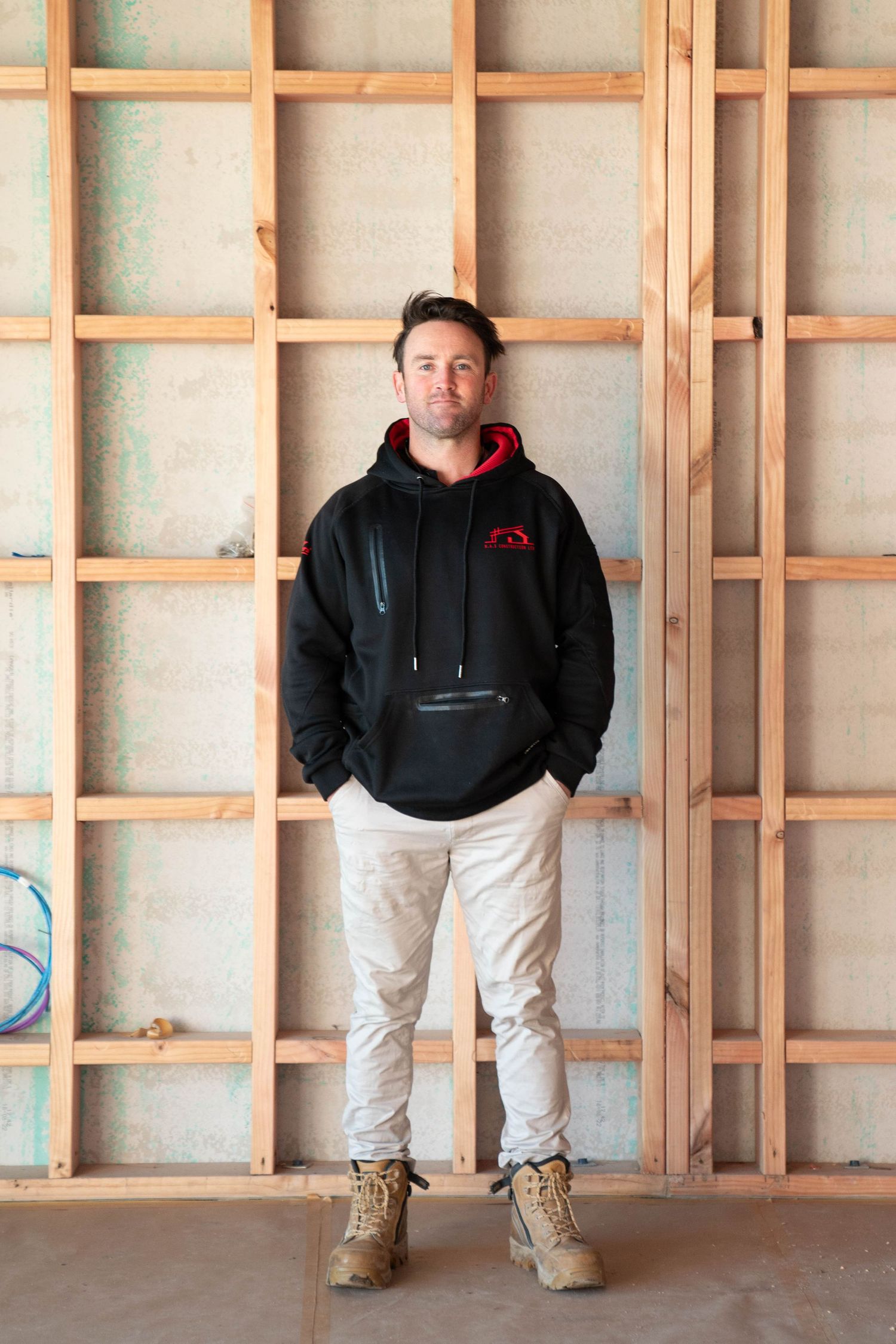 Person wearing black hoodie and tan boots stands against wooden framed wall under construction.