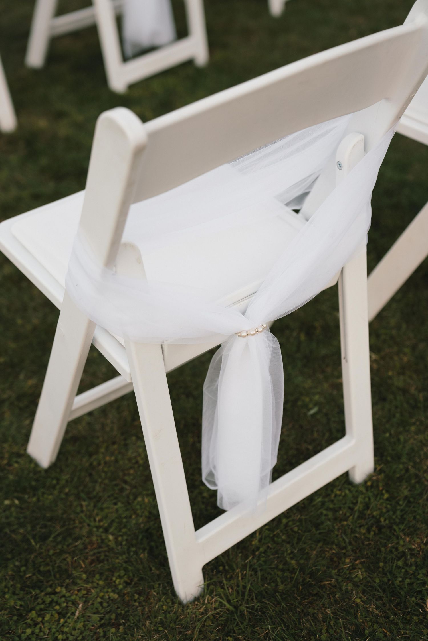 White folding chair with sheer fabric decoration on grass at an outdoor event.
