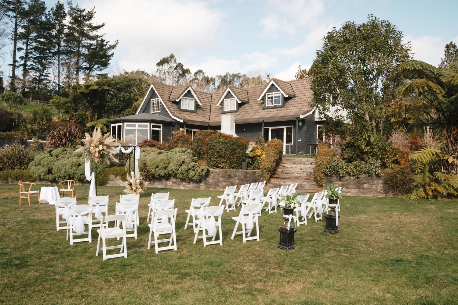 White chairs arranged on a lawn for an outdoor wedding ceremony in front of a charming Cape Cod style home.