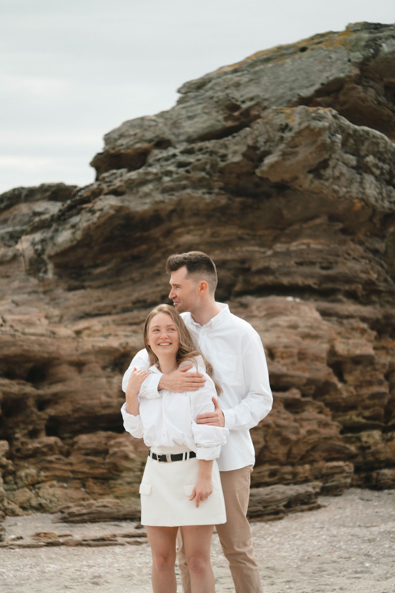 A playful couple shares a tender moment on a beach with rugged rock formations in the background.