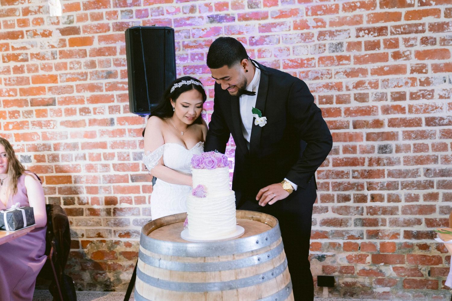 Couple cuts their white wedding cake together on rustic wine barrel display.