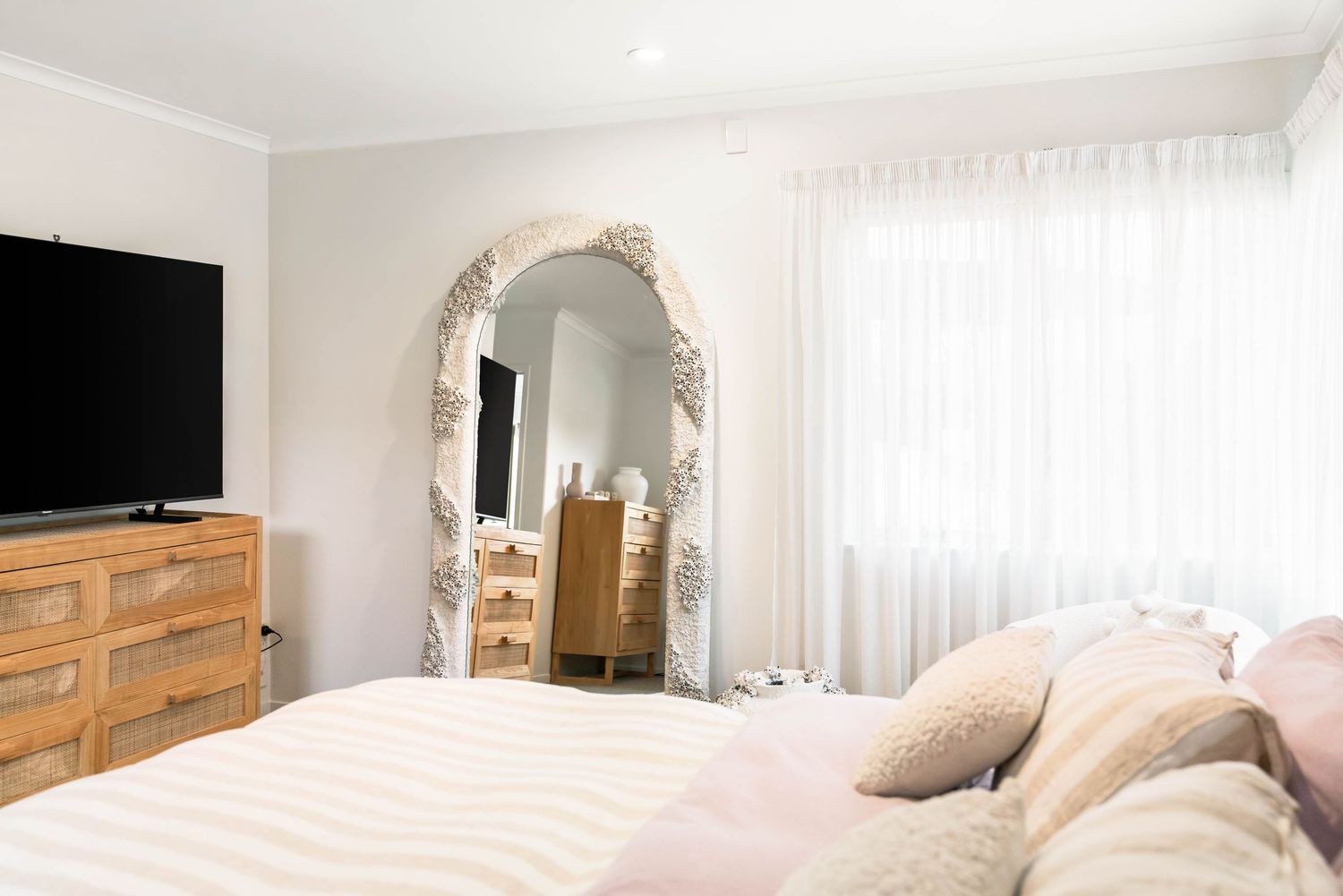 Modern bedroom interior with wooden dresser, ornate white mirror, and natural light flowing through sheer curtains.