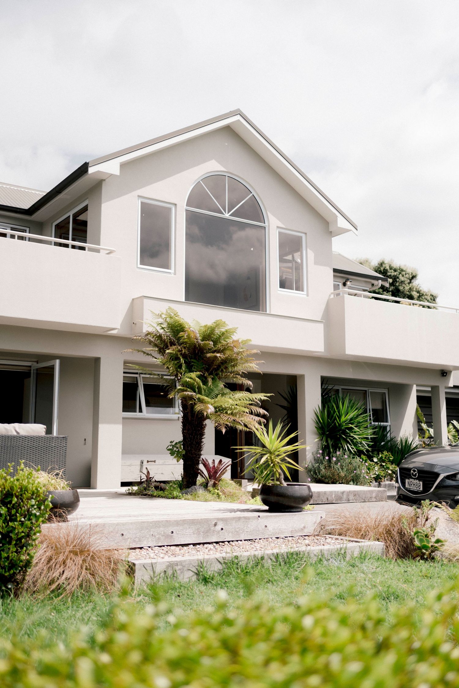 Modern two-story white house with arched window and tropical landscaping in the front garden.