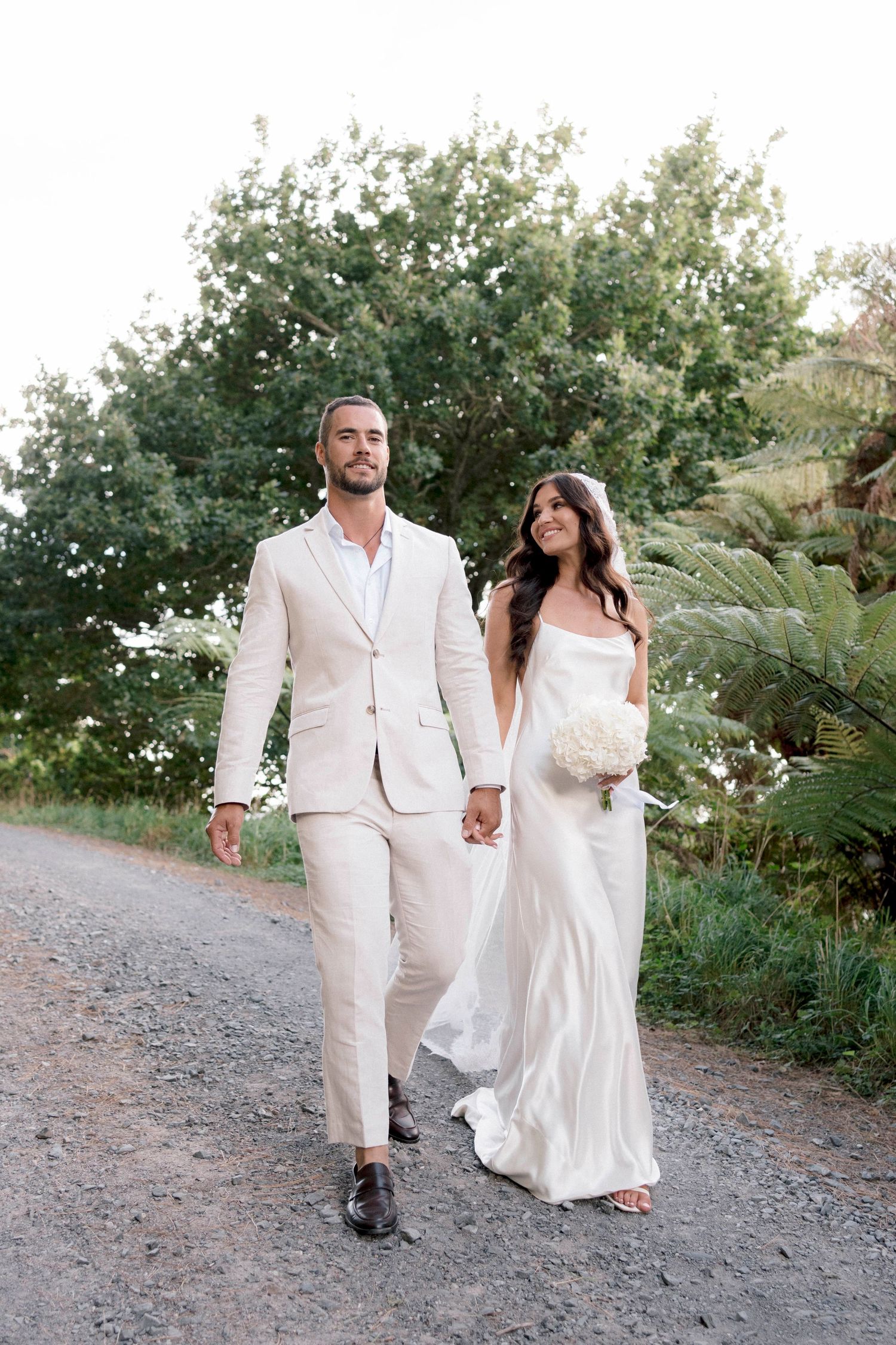 Newlywed couple in cream colored attire holding hands while walking on gravel path surrounded by greenery.