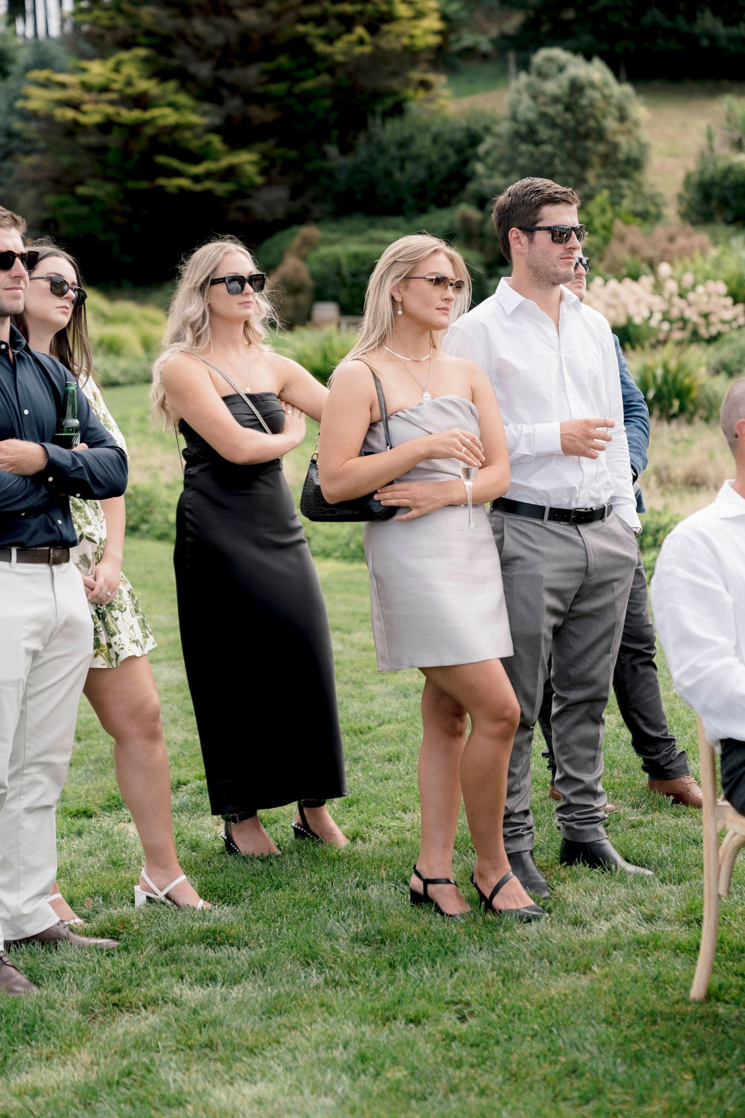 Wedding guests stand in a line on a grassy lawn during an outdoor ceremony.