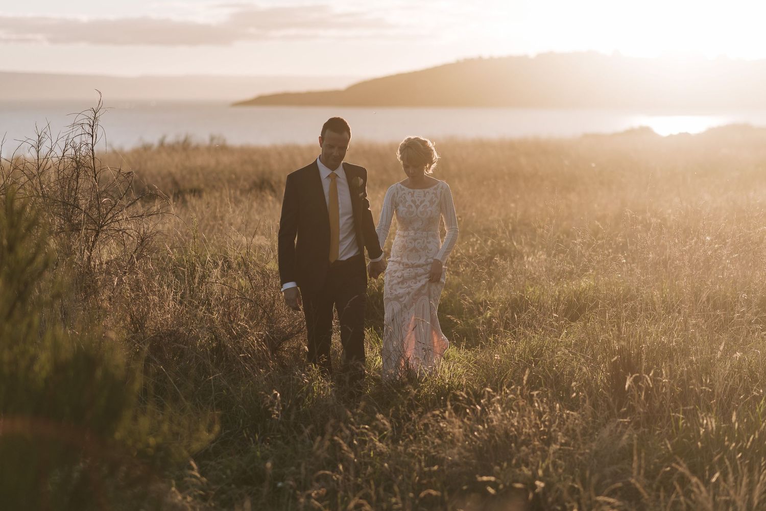 Two figures walk together in a golden field at sunset with mountains in the distance.