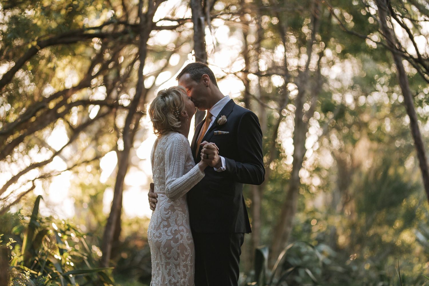 A bride and groom embrace during golden hour in a romantic outdoor forest setting with sunlight filtering through trees.