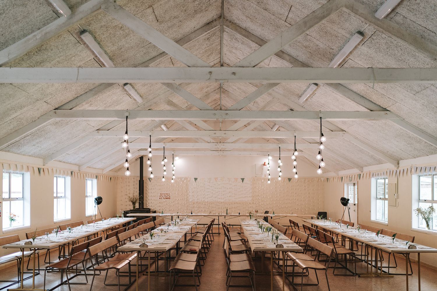 Large rustic dining hall with exposed beam ceiling, track lighting, and long rows of wooden tables in a white interior space.