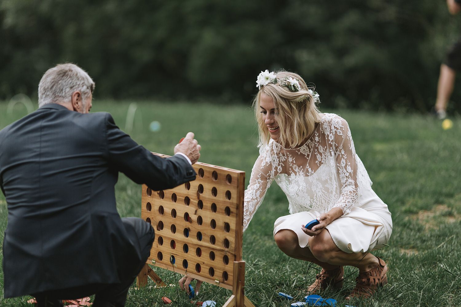 Wedding guests play a large wooden Connect Four game on grass during an outdoor reception celebration.