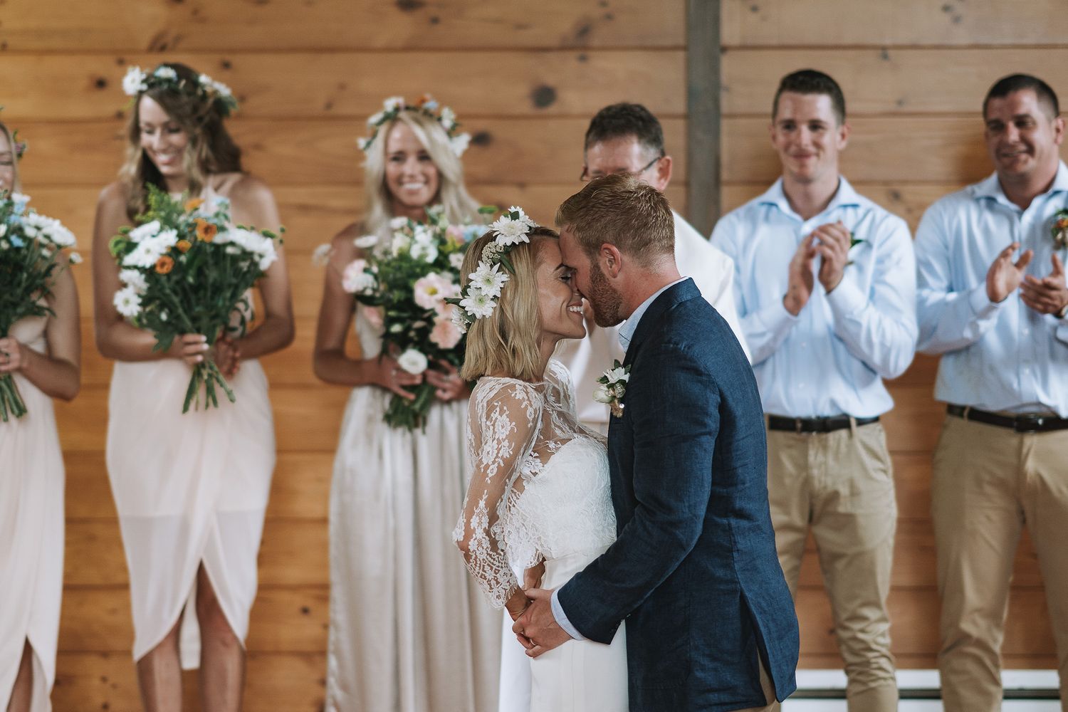 A happy couple shares their first kiss at a rustic barn wedding ceremony with bridesmaids and groomsmen looking on.