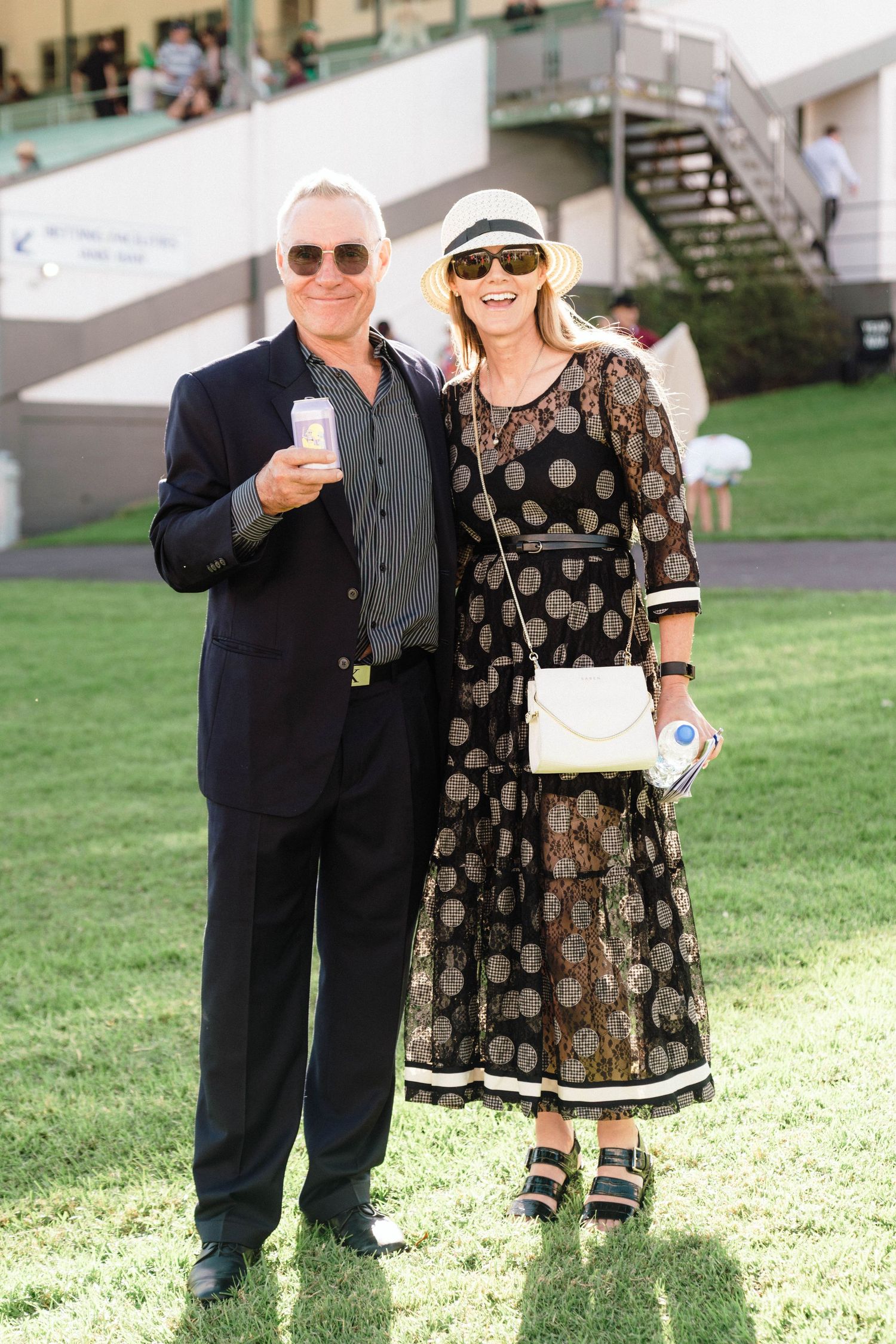 A stylish couple in sunglasses poses on grass at an outdoor event, she wearing a polka dot dress.