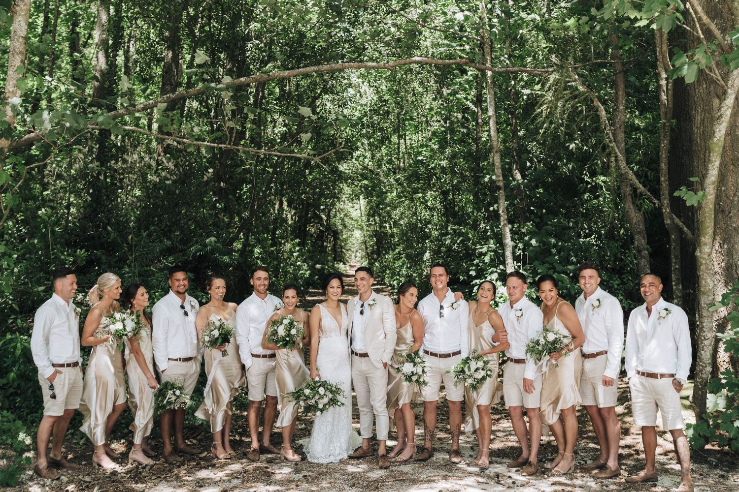 Group of wedding party members in white and beige attire posed together under lush green trees outdoors.