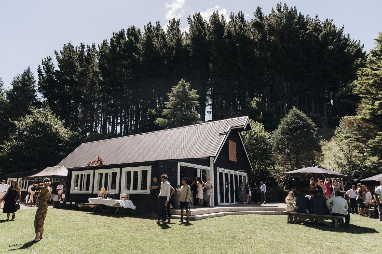 A rustic wooden barn-style building surrounded by tall pine trees with guests gathered outside for an event.
