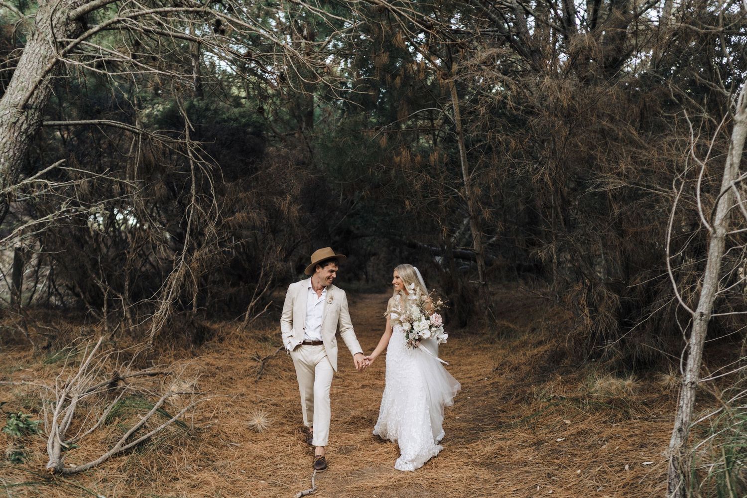 Couple walks hand in hand through wooded trail wearing coordinated white and beige wedding attire.