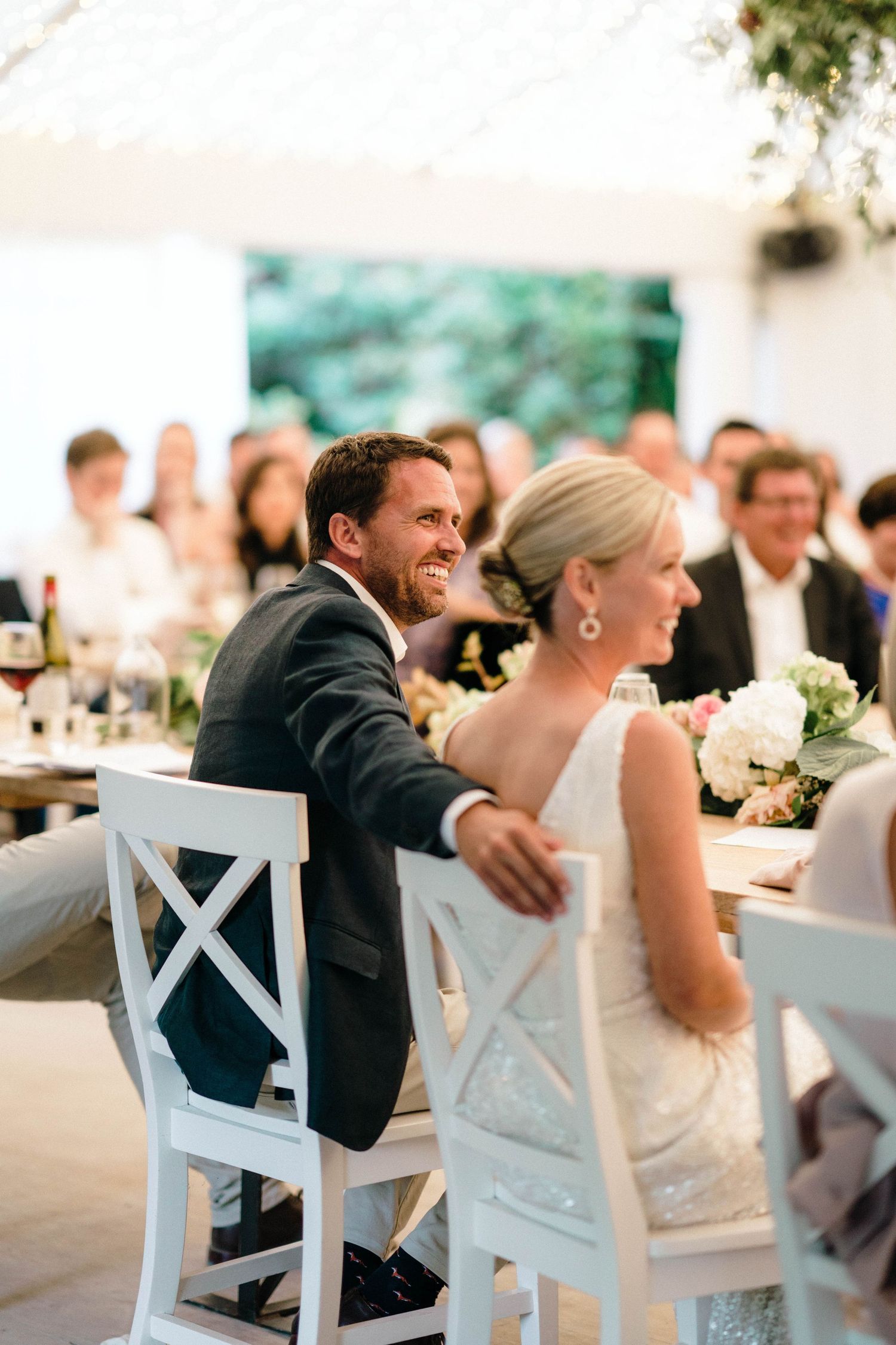 Wedding couple shares candid laughter during reception dinner at white cross-back chairs.