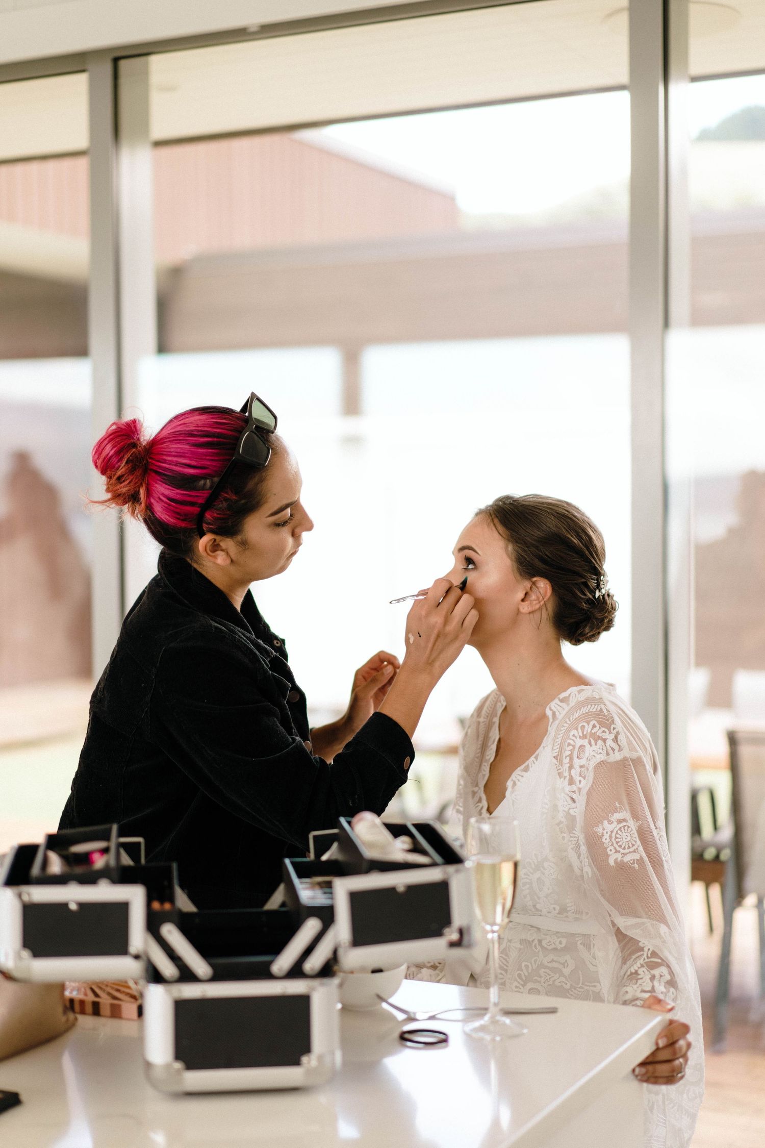 Makeup artist applying cosmetics to a person wearing a white lace dress in a bright indoor setting.