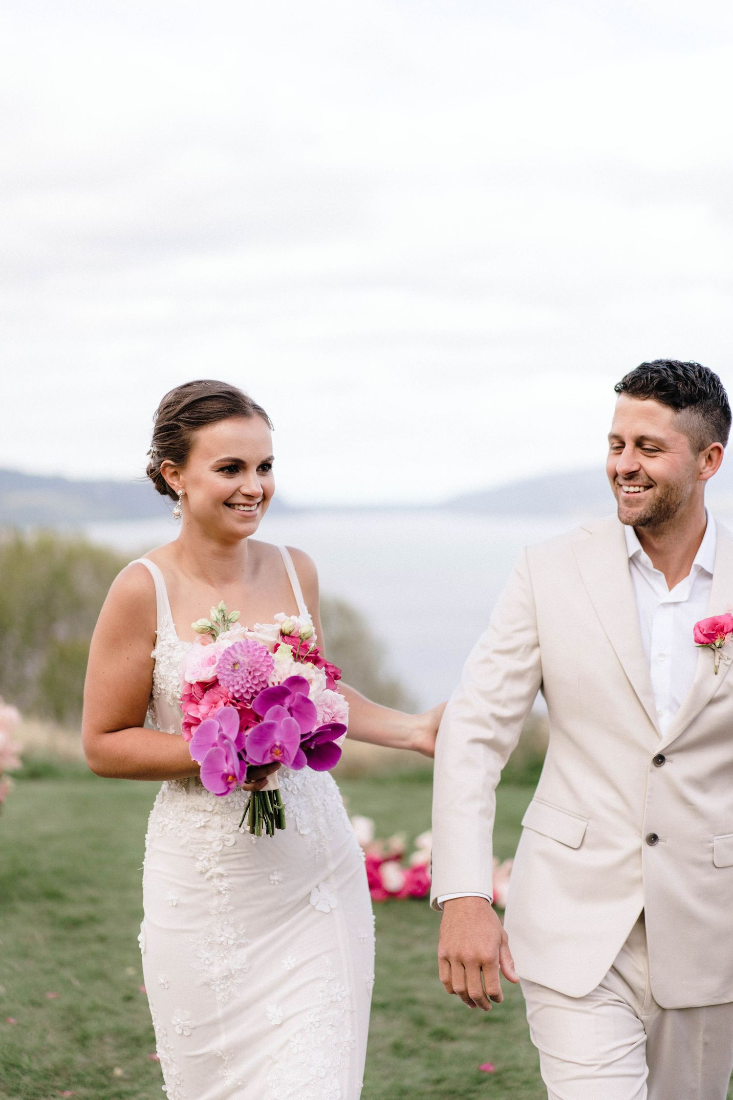 Newlyweds in white attire walking and laughing together on grassy hillside with pink bouquet.