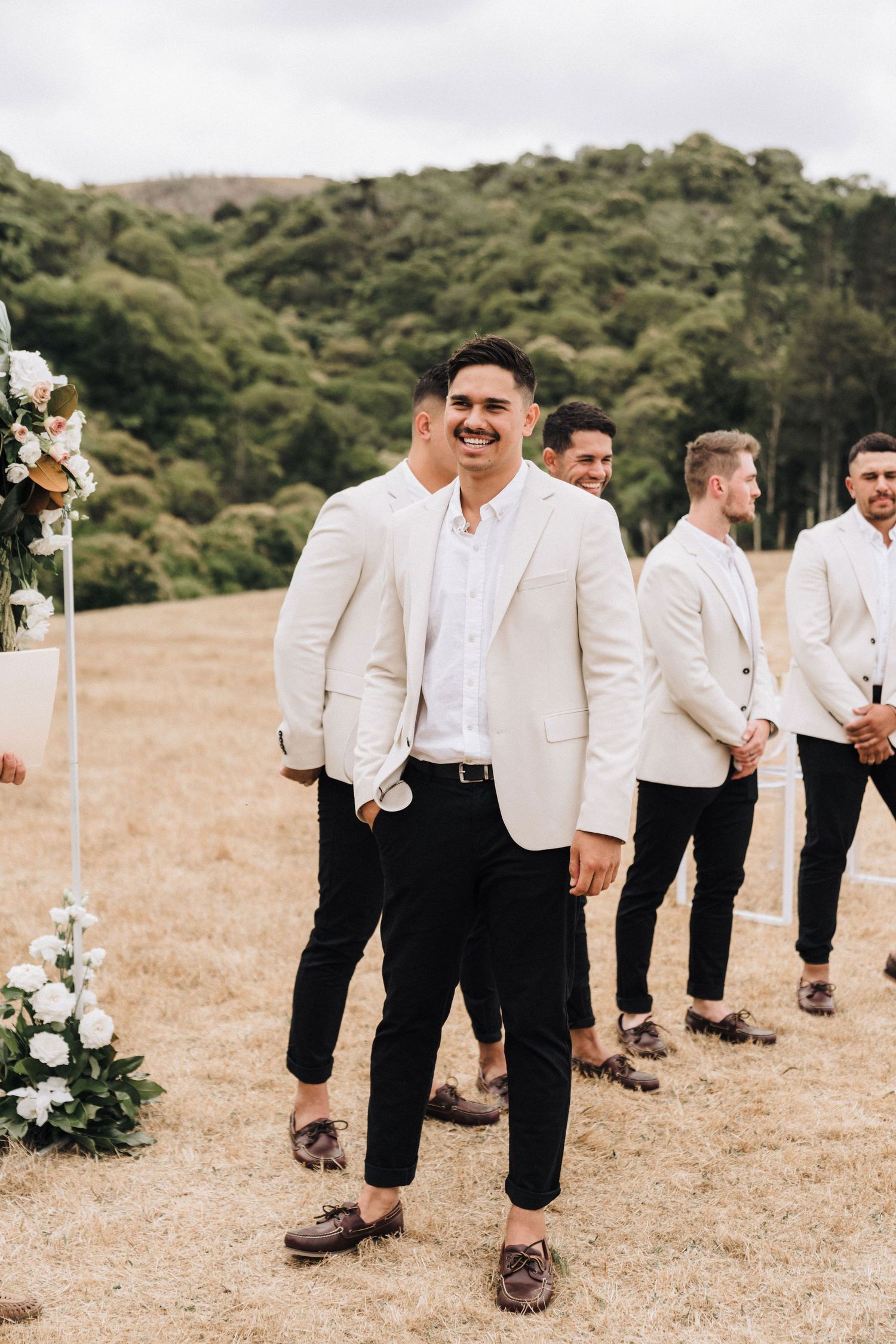 Groomsmen in white jackets and black pants walk across a sunlit field during an outdoor wedding.
