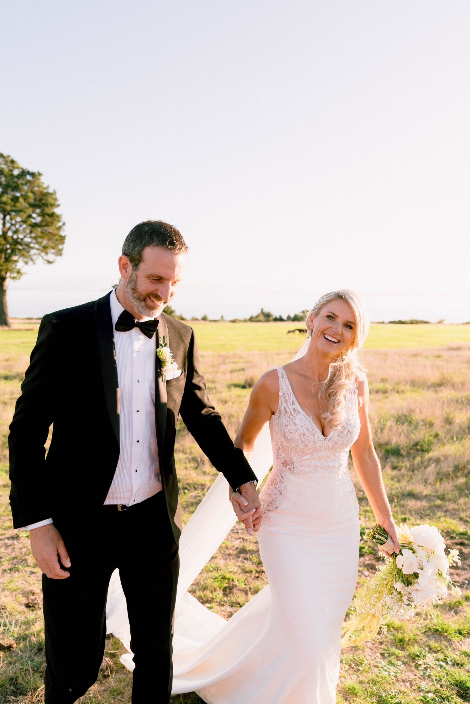 A joyful couple in wedding attire hold hands while walking through a sunlit field at golden hour.