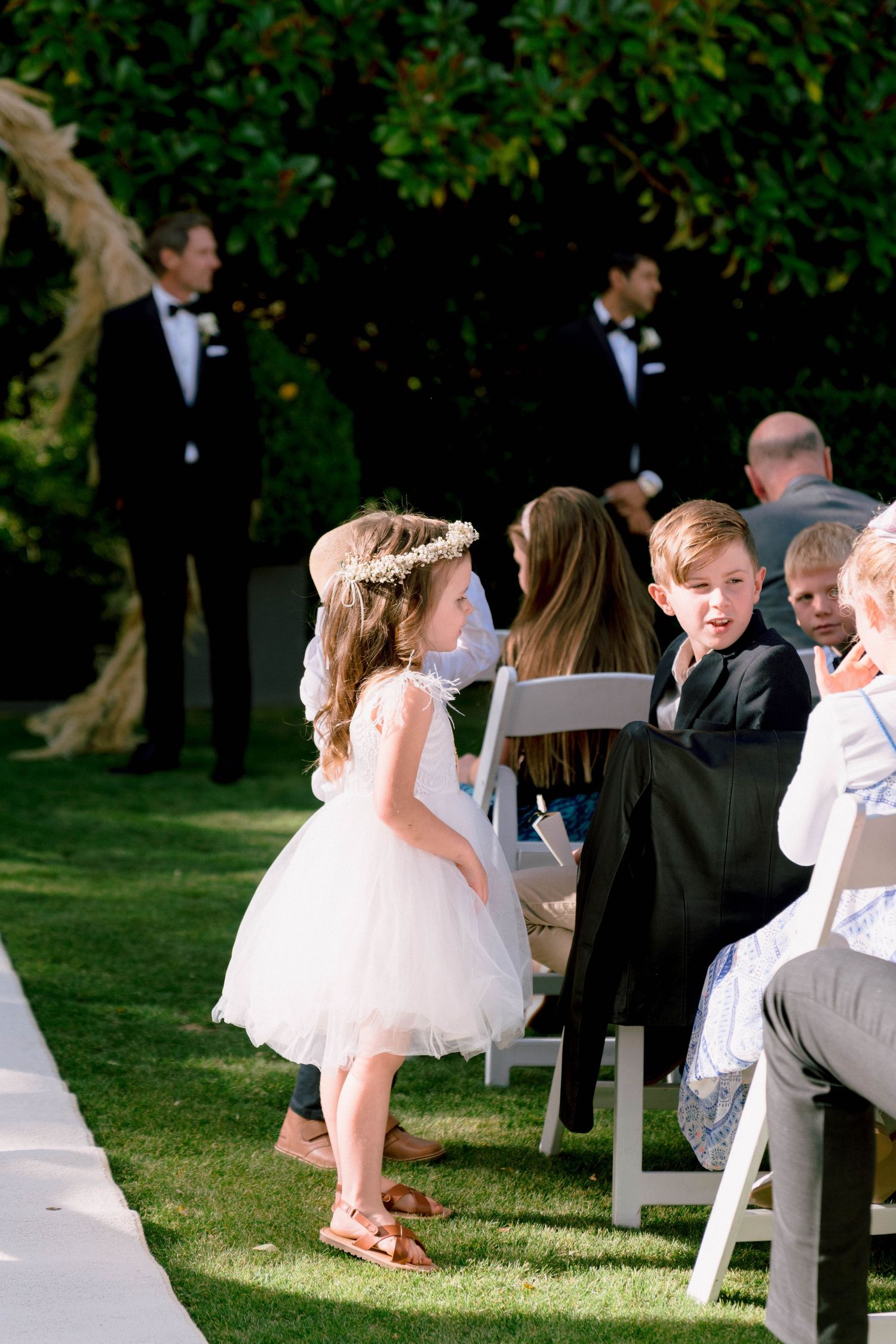 Wedding guests seated on white chairs outdoors during a ceremony with lush greenery in the background.