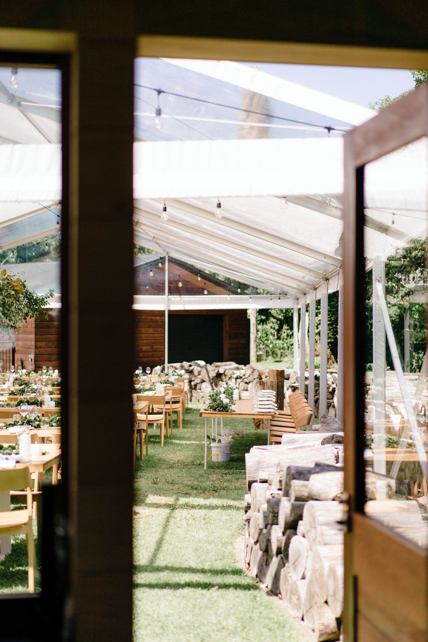 Outdoor wedding reception tent setup with wooden tables and natural lighting over grassy lawn.