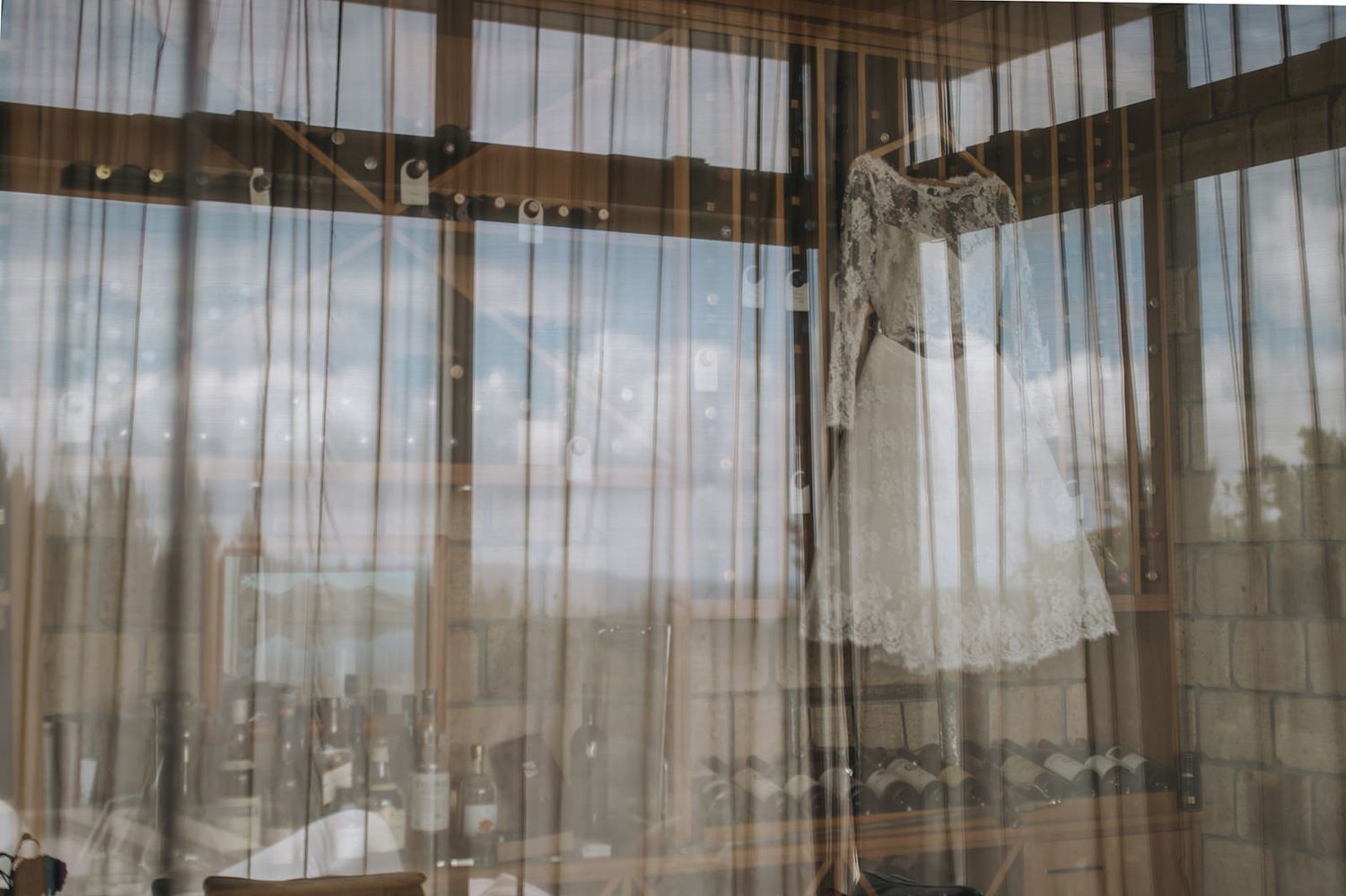 A white wedding dress hangs from a curtain rod in front of sheer window drapes with sunlight streaming through.