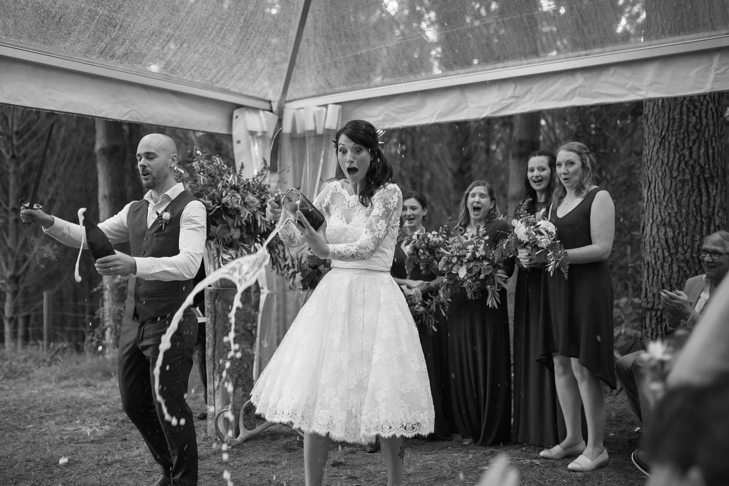 A couple celebrates during an outdoor wedding ceremony under trees while bridesmaids in dark dresses look on.
