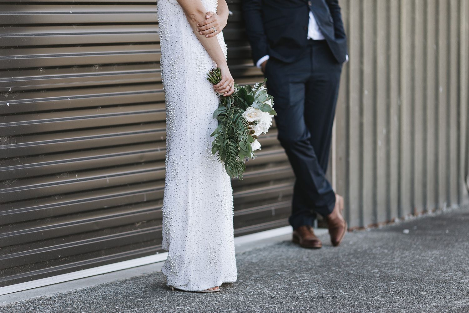 A couple in wedding attire stands against an industrial wall, with the bride holding a simple green bouquet.