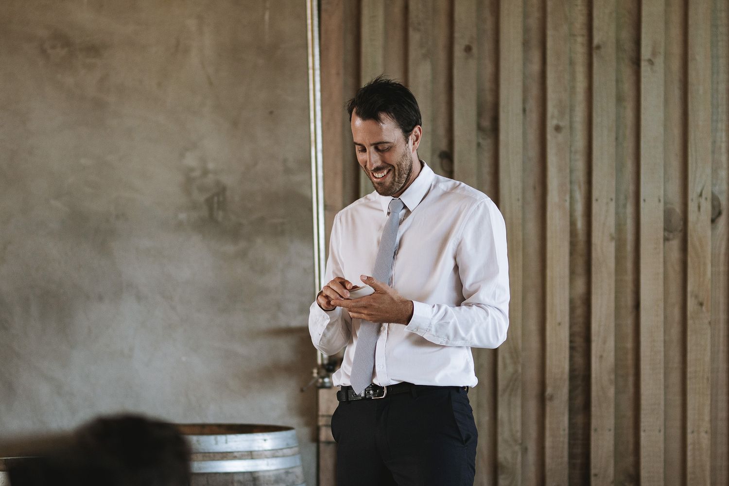 A person in a white shirt stands indoors against a wooden wall backdrop textures, looking at a phone with a smile.