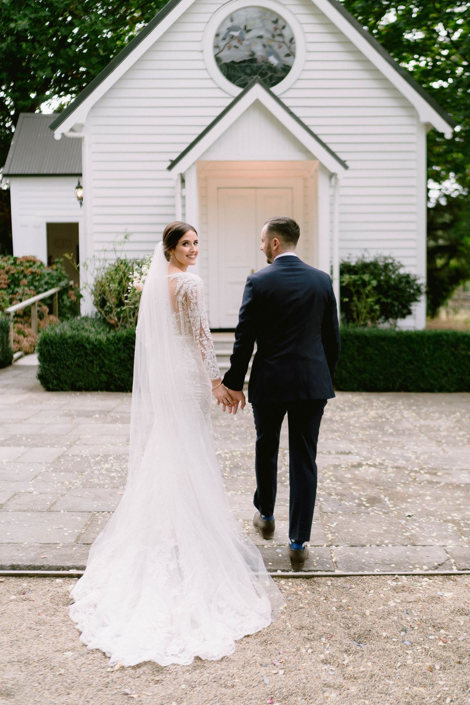 A couple approaches a charming white chapel with a circular window on their wedding day.
