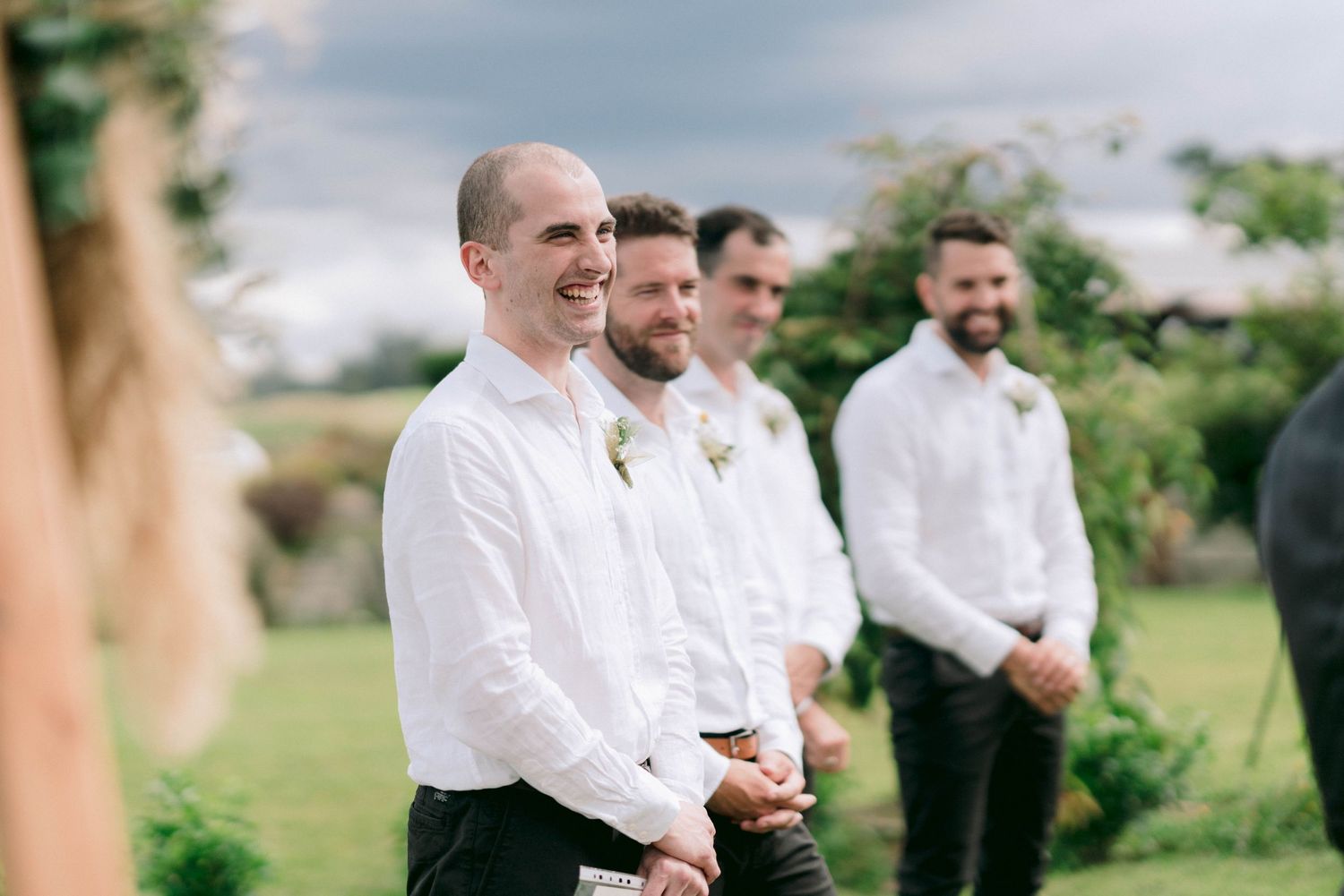 Groomsmen in white shirts and dark pants standing in a line outdoors during wedding ceremony.