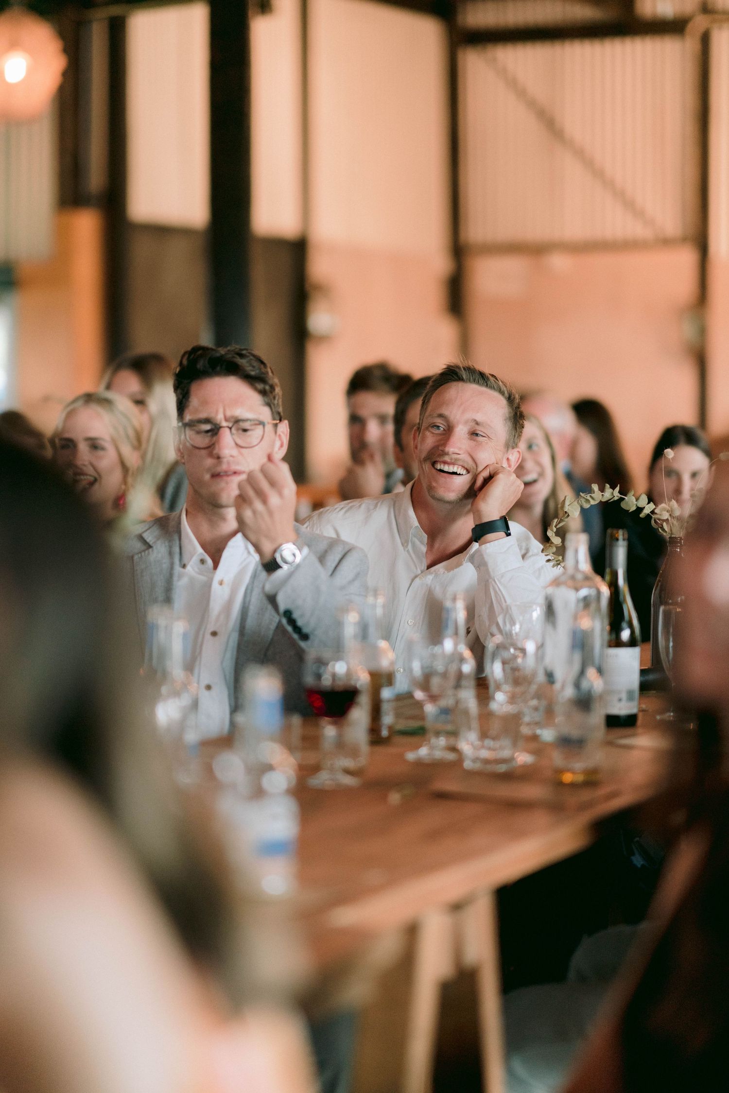 Wedding reception guests laughing while seated at wooden tables with wine glasses.