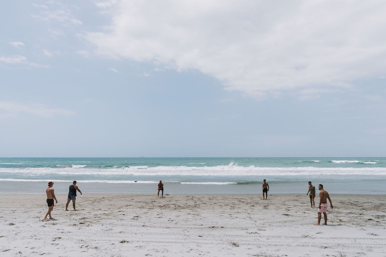 Silhouettes of beachgoers walking along the sandy shoreline as gentle waves roll in under a cloudy coastal sky.