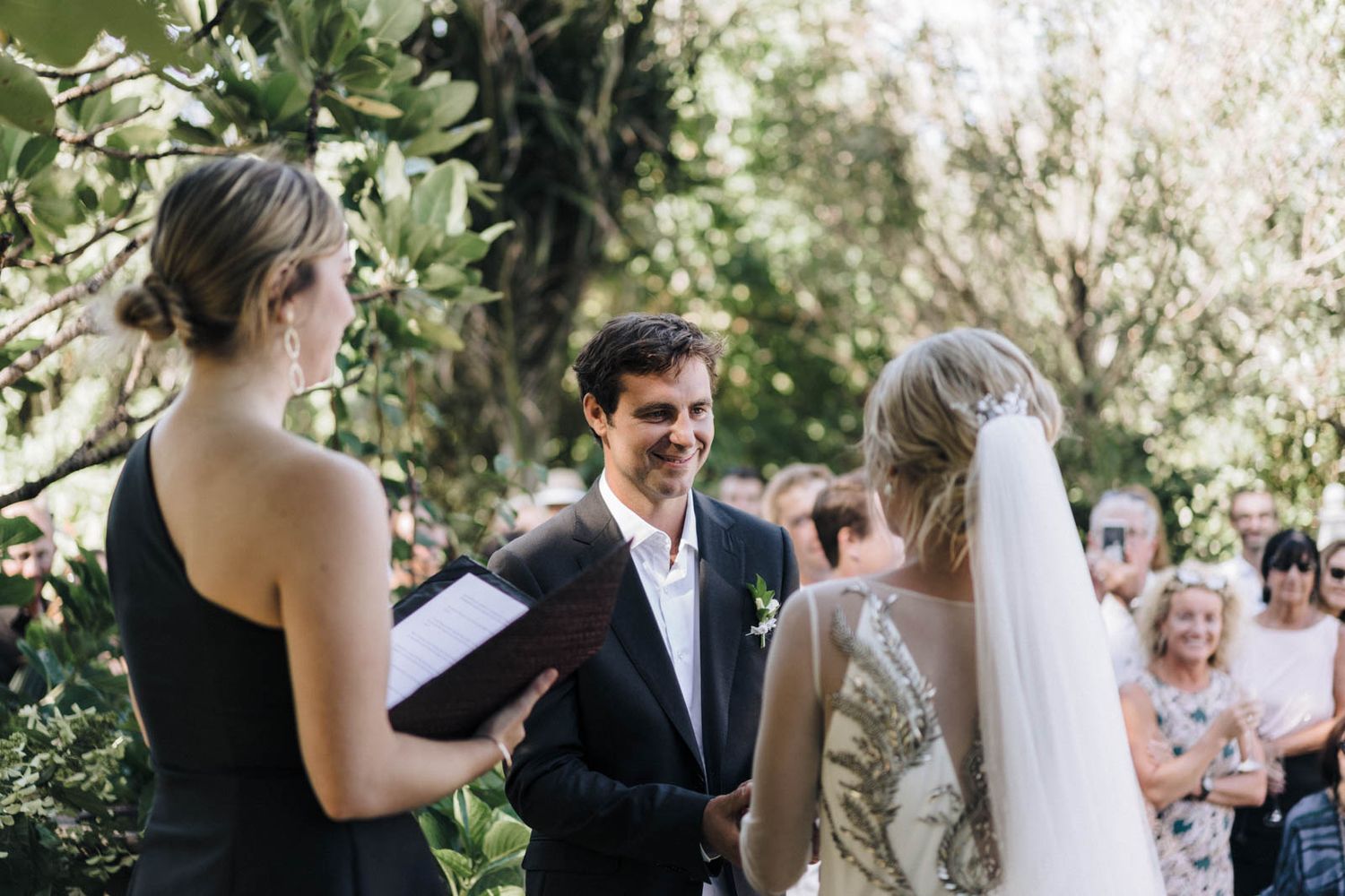 Wedding guests celebrate and applaud during an outdoor garden ceremony on a sunny afternoon.