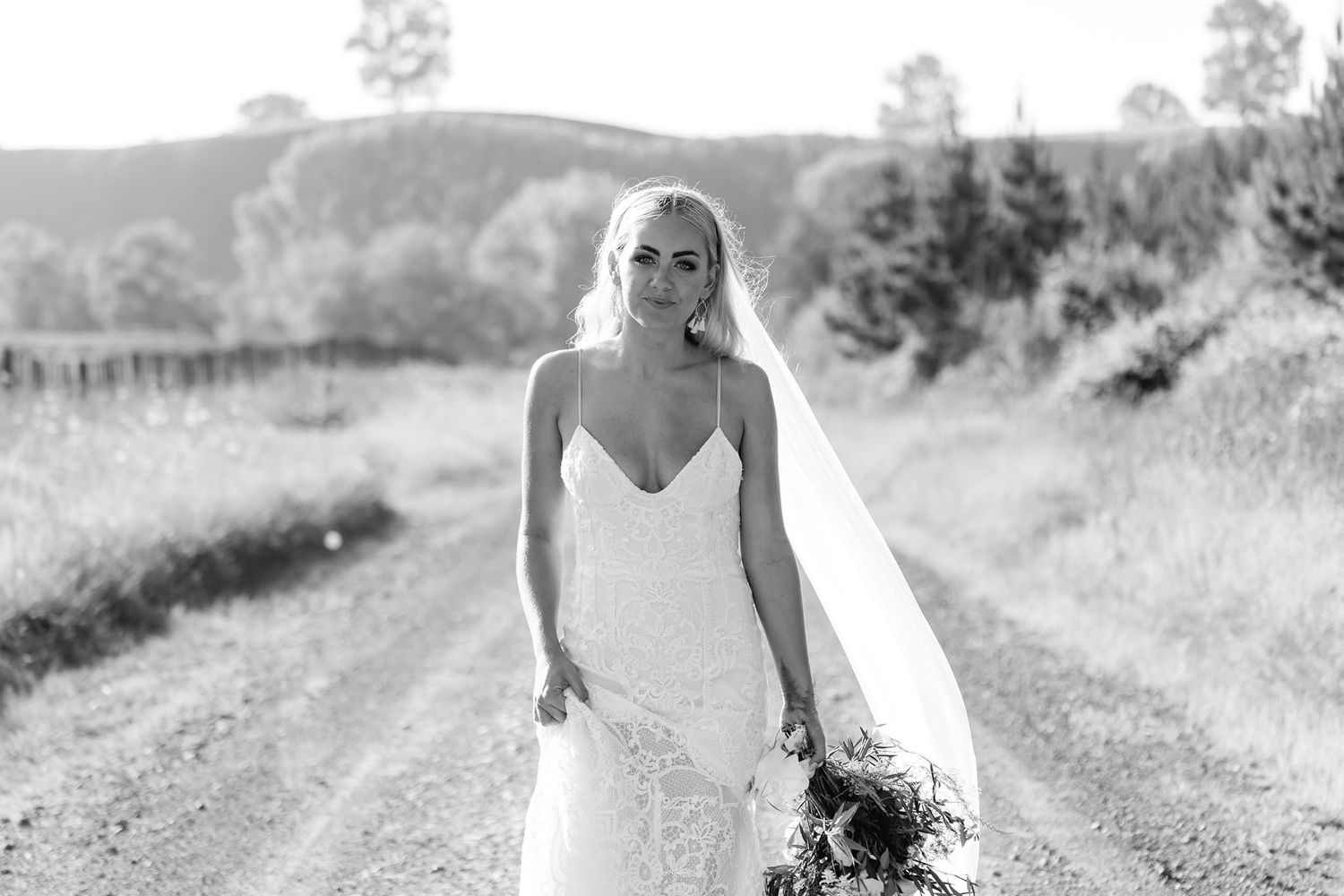 A bride in a sweetheart neckline gown stands on a rural dirt path holding her bouquet in black and white photography.