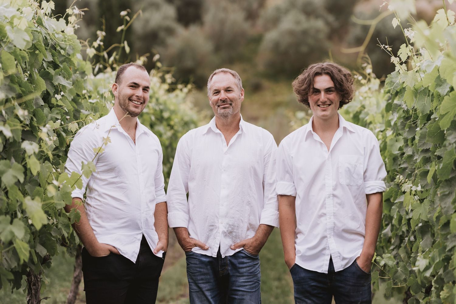 Siblings wearing coordinating white shirts pose casually together for family portraits in a lush green vineyard.