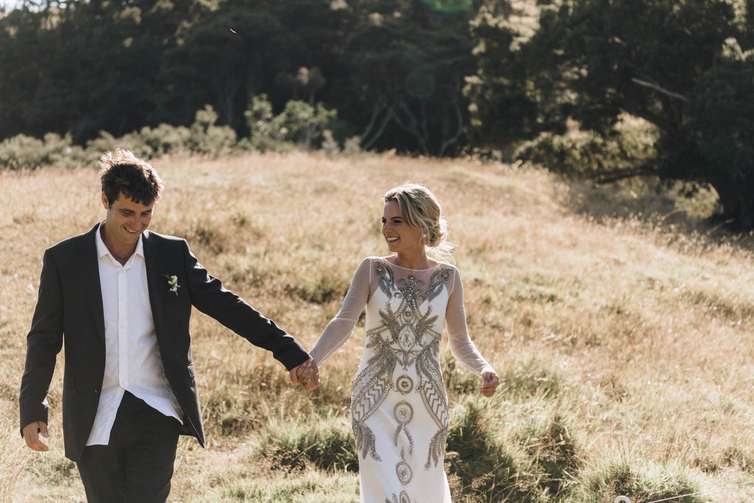 A couple walks through a sunlit field wearing formal attire against a scenic natural backdrop.