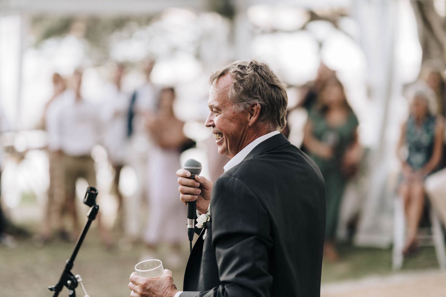 A person addresses wedding guests at an outdoor reception while holding a microphone and champagne glass.