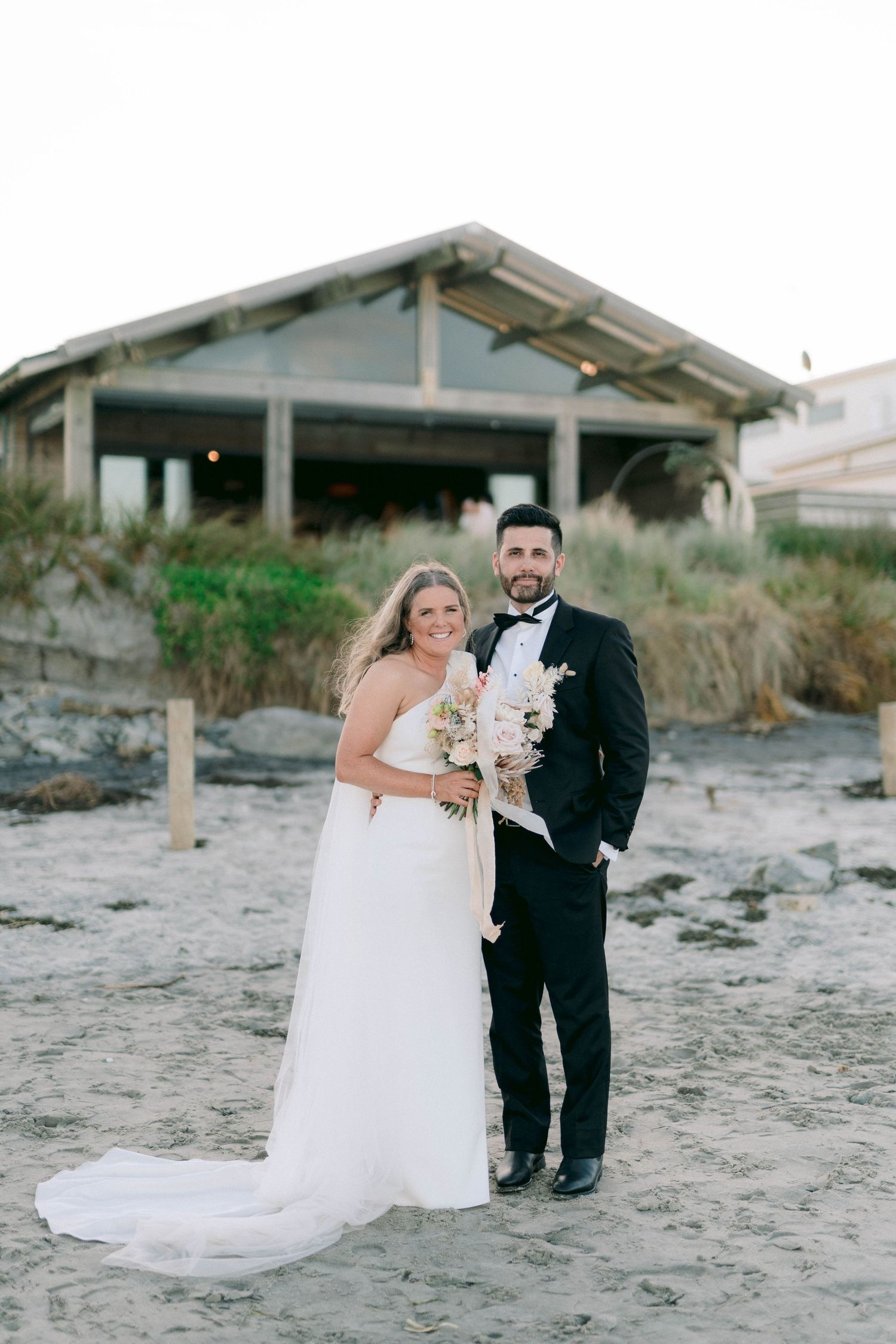 Elegant beach wedding couple poses on sandy shore with rustic building backdrop and white bouquet at sunset.