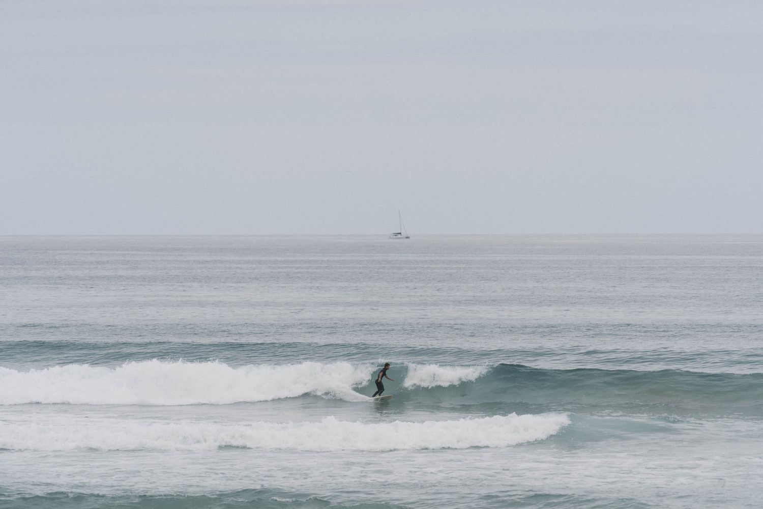 A lone surfer rides a turquoise wave in a misty gray seascape with a ship visible on the horizon.