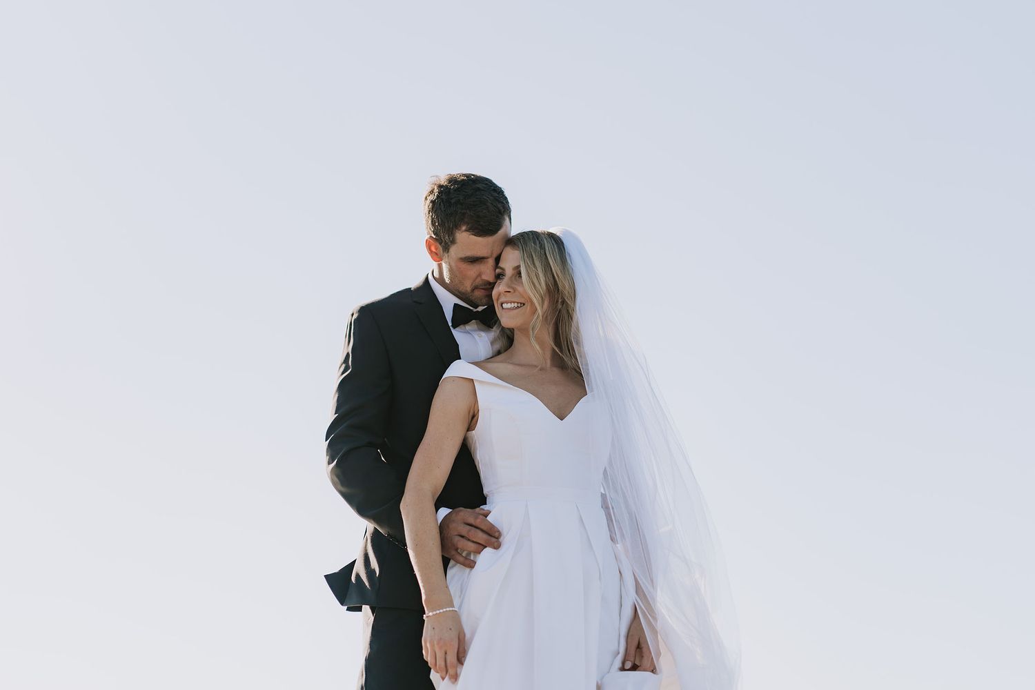 A bride in a white wedding dress and groom in dark suit embrace against a light sky background.