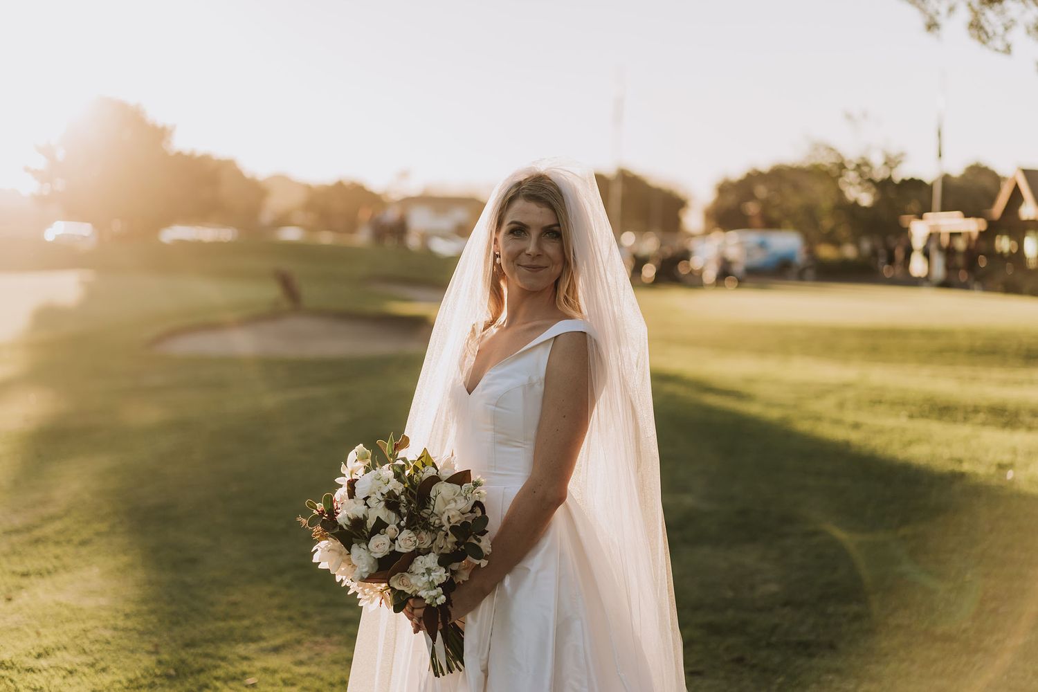 A bride in a white wedding dress holds a beautiful bouquet of flowers during golden hour at sunset on a golf course lawn.