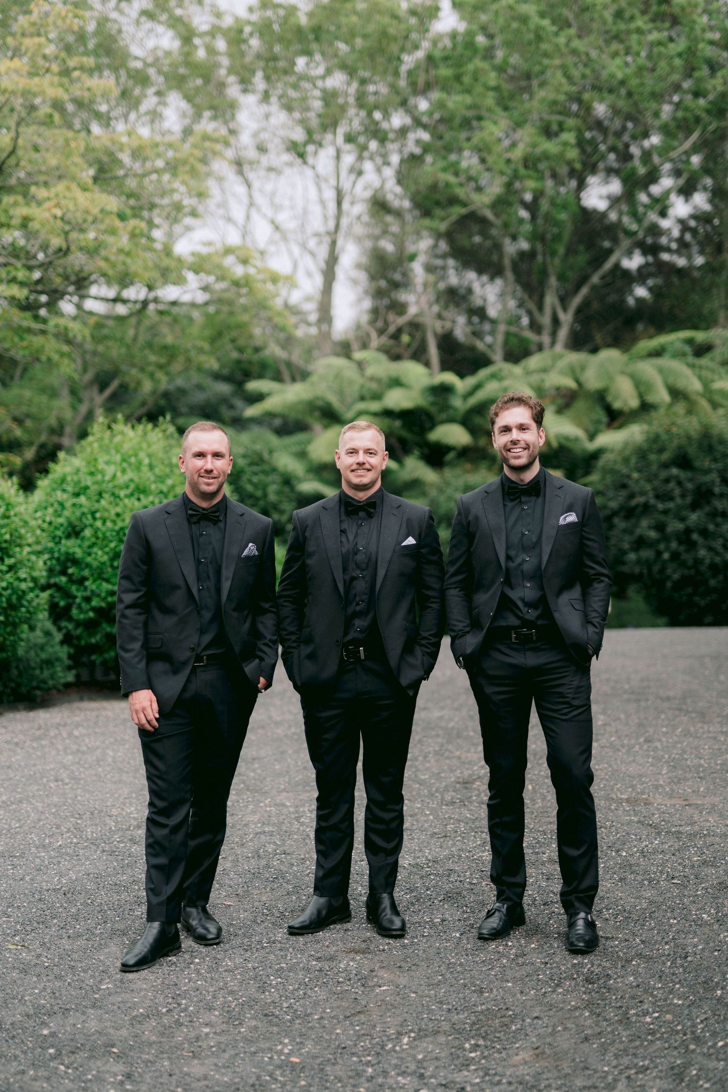 Three groomsmen in matching black suits pose for a photo on a garden path.