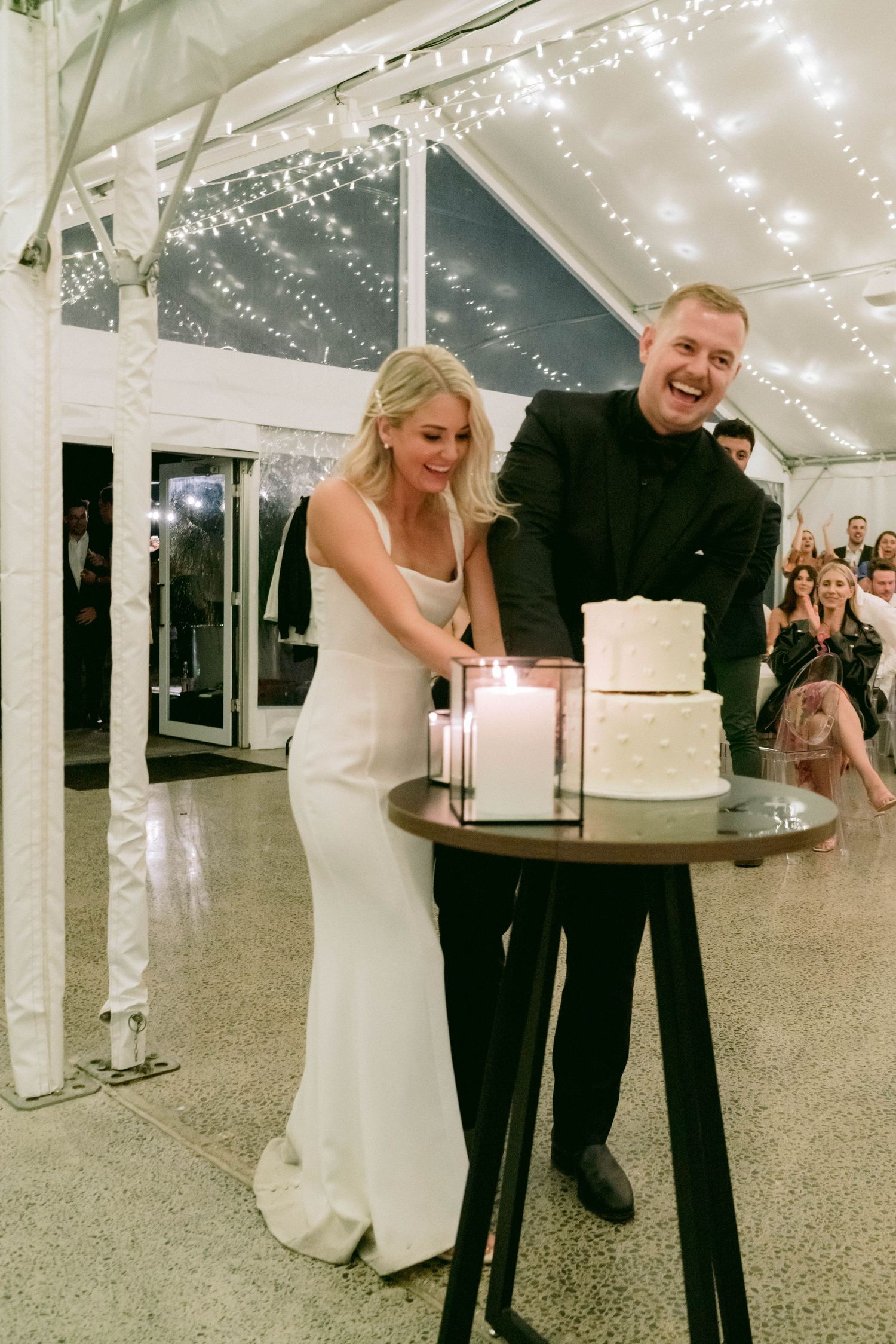 A couple cuts their wedding cake together under twinkling lights at their reception venue.