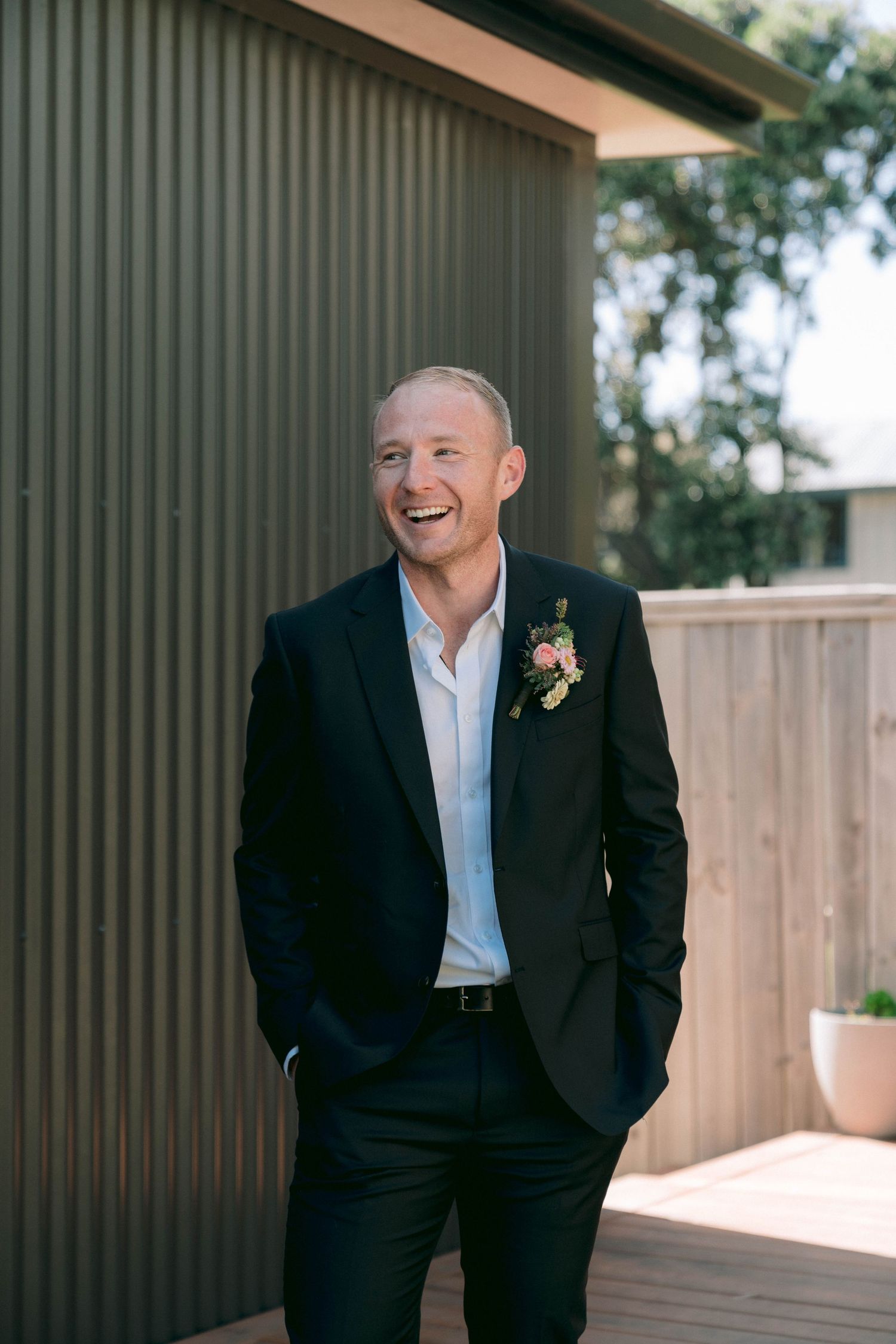 A groom in a black suit and white shirt smiles joyfully while standing against a wooden wall outdoors.