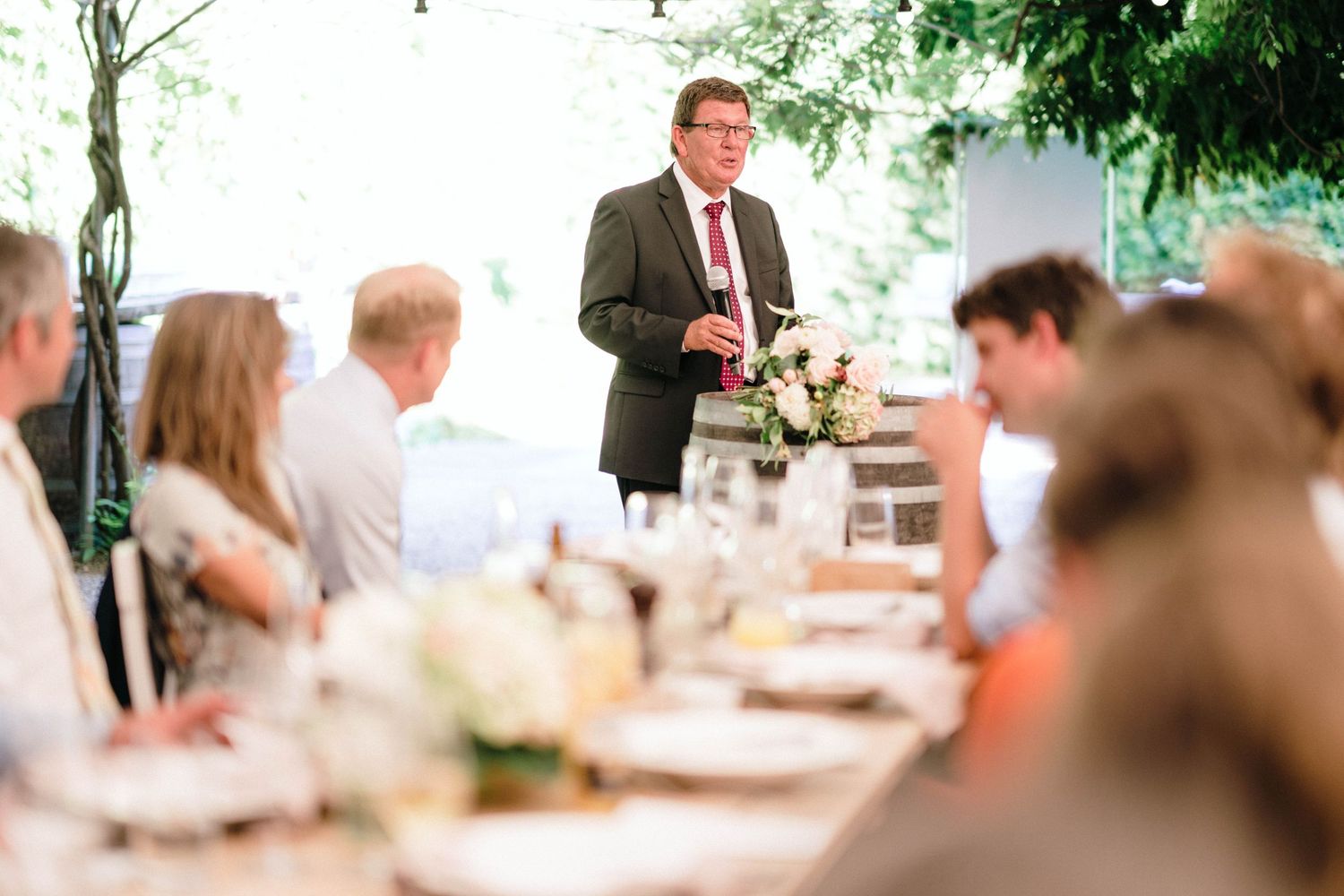 Blurred reception dinner scene with green foliage and warm lighting overhead.