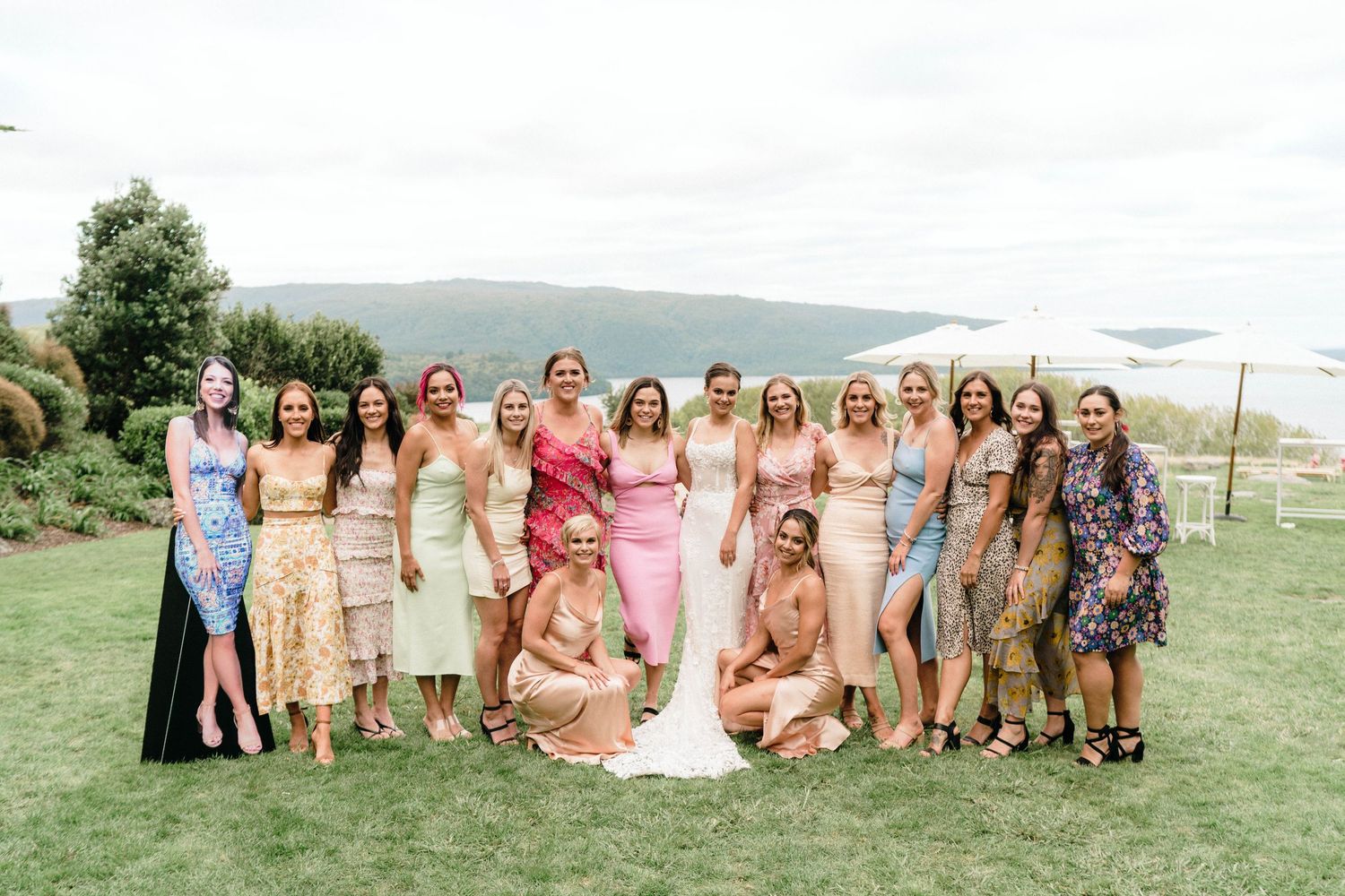 Large group of wedding guests in pastel dresses posing on grassy hillside.