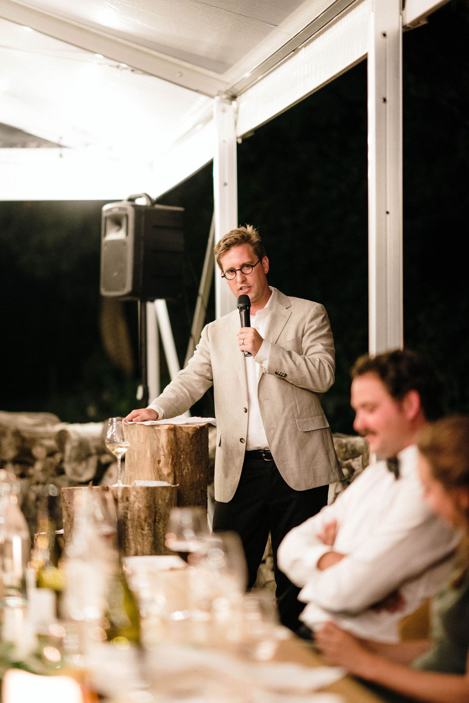 A speaker in a light colored suit gives a speech at an evening reception with table settings visible in the foreground.