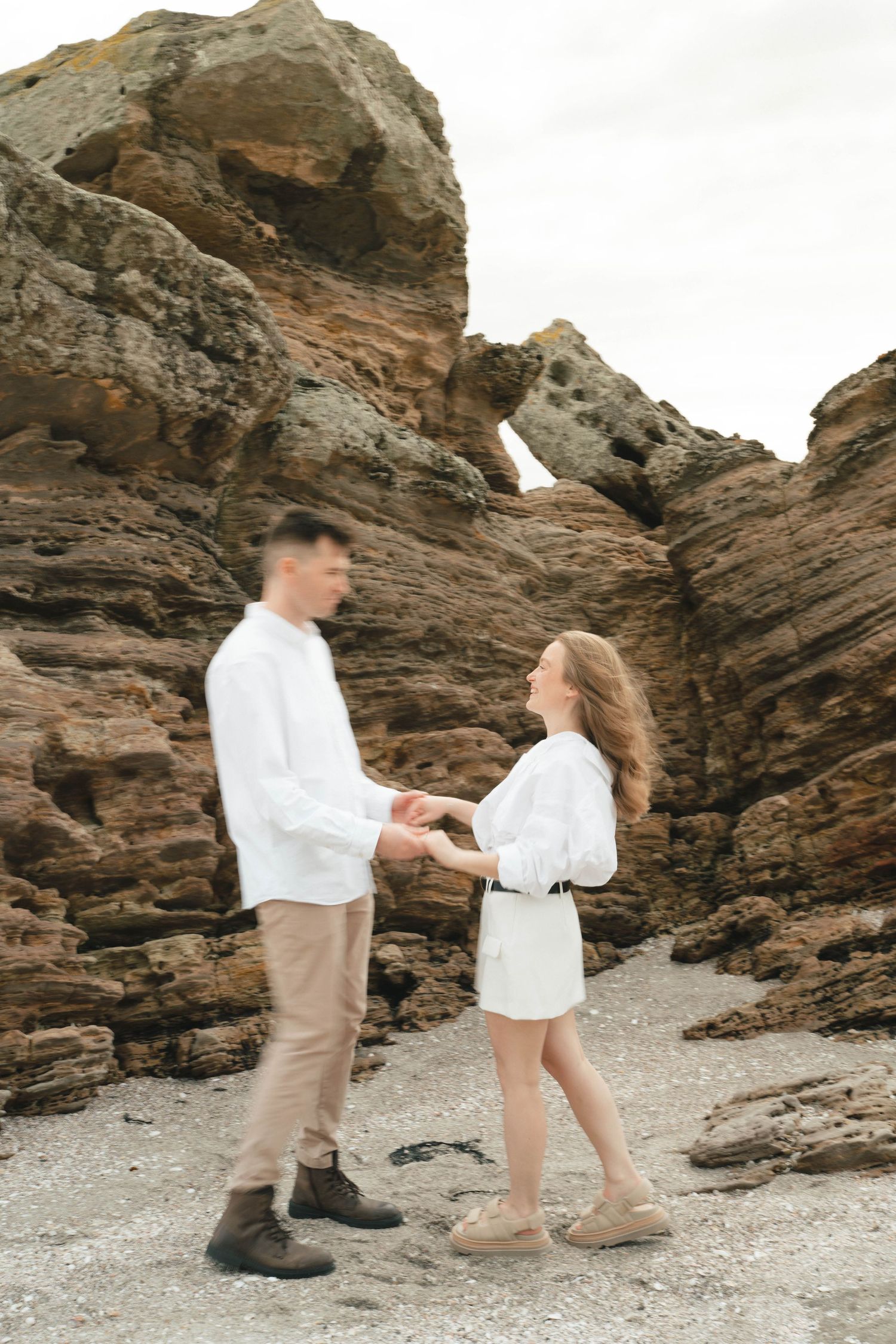 A couple in white clothing dances playfully on a rocky beach surrounded by cliffs.